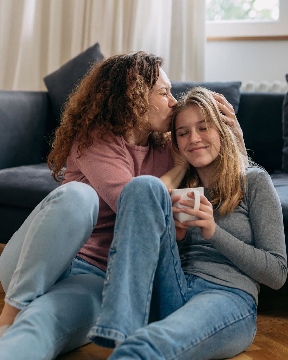 Mother kissing the side of her teenage daughter's head while the two of them sit on the floor together