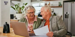 Smiling senior couple discussing bills and reviewing documents while using laptop at home. Mature couple collaborating on financial management at their dining table. Elder man and retired woman sharing a moment while organizing their finances while using computer.
